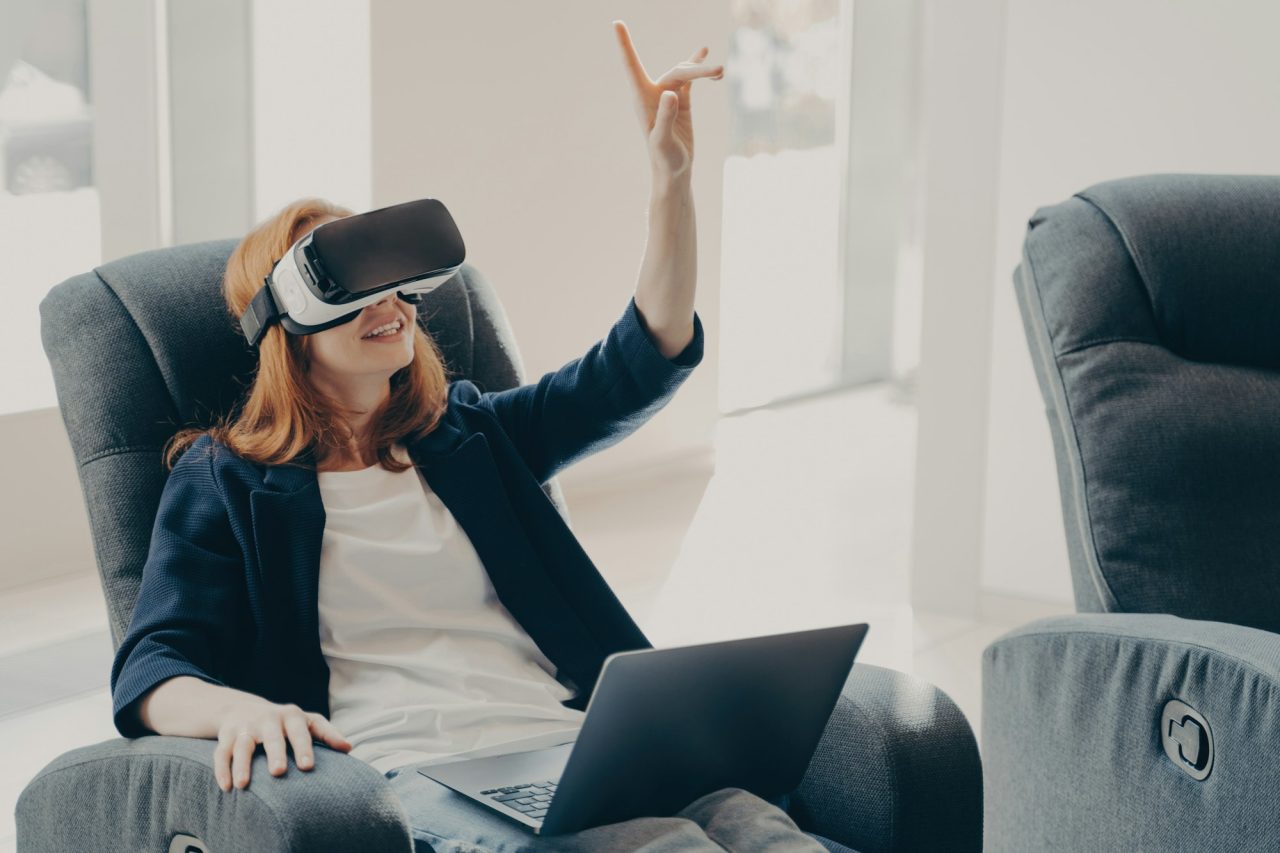 female in vr headset pointing with finger touching 3d object while working remotely on laptop